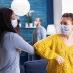 cheerful-african-woman-her-friend-touching-elbow-keeping-social-distancing-while-greating-each-other-wearing-face-mask-prevent-coronavirus-spread-course-global-pandemic-living-roo-min